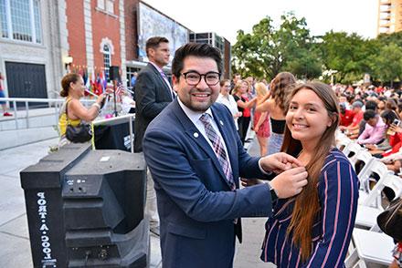 2019 alumni board president pinning lapel pin on incoming female student both looking at camera with smile in front of fine arts building